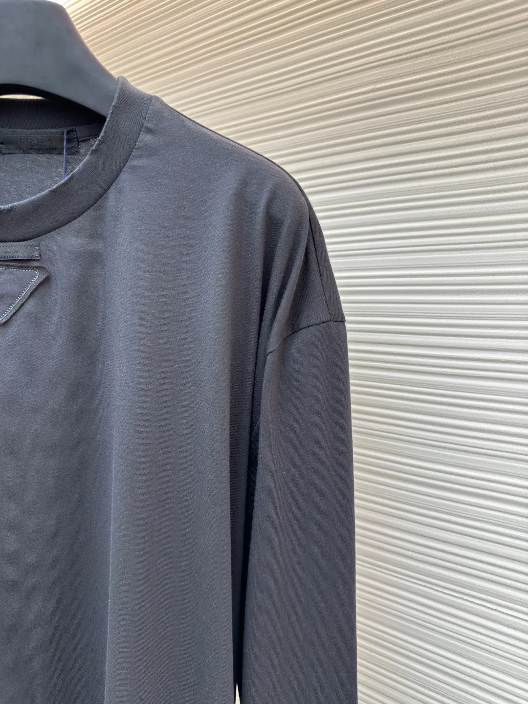 Prada Pencil Gray Long Sleeve ●Original version: Purchased and developed by 8934 ●Item number: 1680720-A05 ●Fabric: combed plain weave fabric ●Technology: Laser burning ●Version: fitted version ●Weight: about 240 grams ●Size: S-M-L-XL ●Description: Custom-woven 50S 190G combed plain weave fabric, non-market general gray fabric, needs to be fully customized from yarn ratio and loom parameters. Short fiber impurities are removed through the combing process to ensure that the yarn is longer and more uniform, and the fabric surface is smooth and hairless.  Specially dyed pencil gray, the color is adjusted individually instead of using a standard color card. Multiple proofing is required to match the low saturation gray tone specified by Prada. Chest triangle laser burnt embroidery shaping: Laser burnt: high-precision laser engraving, carbonizing the fabric surface to form a gradient etching texture, and the edges need to be manually adjusted to avoid scorch marks.  Composite embroidery: superimpose embroidery on the basis of burnt flowers. The stitch density needs to be aligned with the burnt flowers pattern to prevent fabric deformation.  Three-dimensional shaping: Use high-temperature pressing or resin coating to maintain the relief of the triangular structure and avoid collapse after washing. The overall wiring is straight and delicate, the fabric is comfortable and soft, and the color is consistent without color difference. It can be matched with the same style to create a fashionable look and control various occasions. Customized accessories, complete with three standards, same style for men and women!