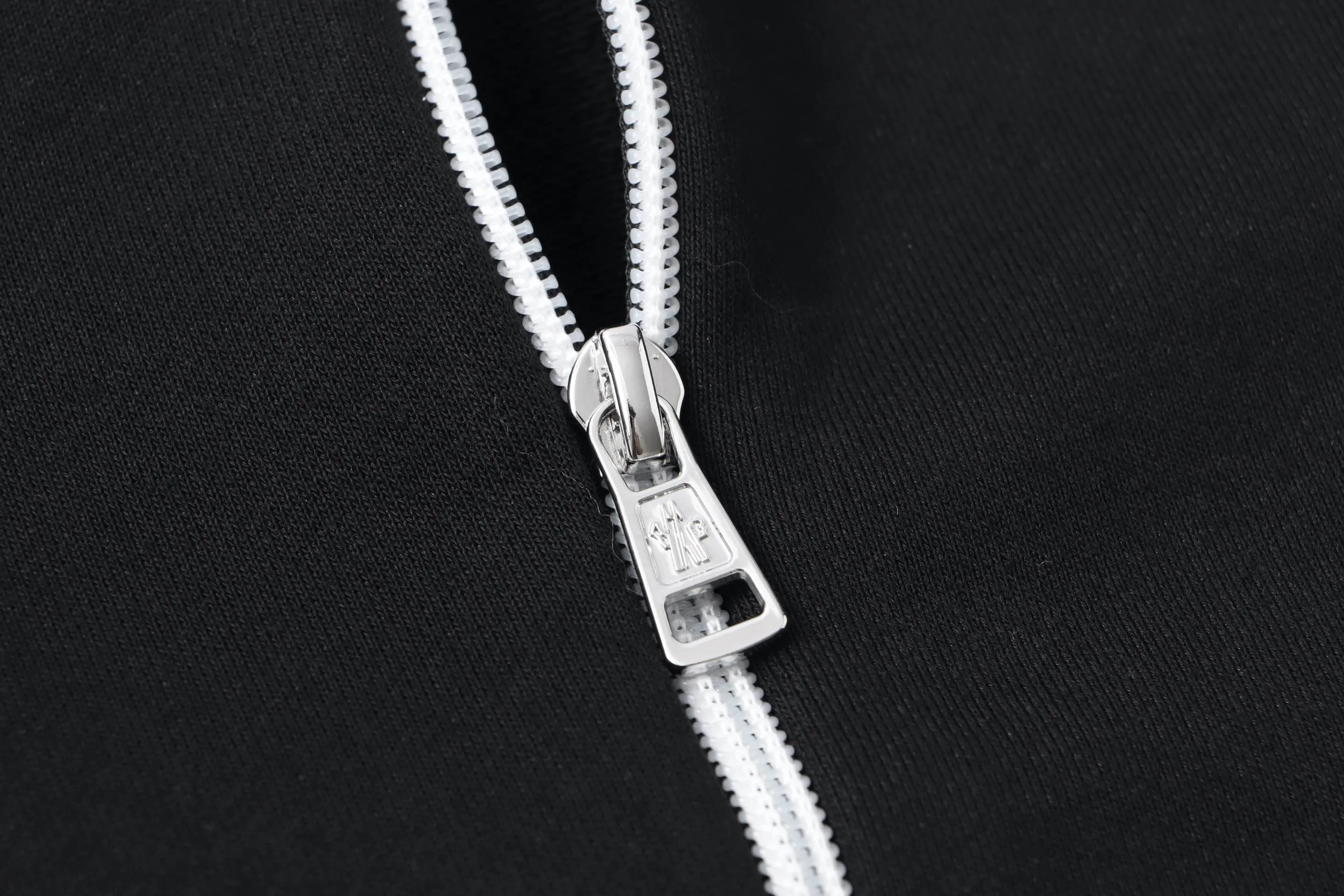 P340 Moncler slogan printed zipper hooded jacket is made of lightweight turned velvet cotton fabric. High-density cotton fiber is used to achieve internal and external differentiation in a single layer of fabric. The outer plain weave is tight and wear-resistant, and the inner layer is sanded to form a short dense suede surface. The dense plain weave structure resists wind and splashes, the brushed suede surface is skin-friendly, and the short pile has been treated to eliminate static electricity to avoid sticking to the skin. A very small amount of grooved polyester moisture-conducting yarn is embedded in the cotton fiber, which quickly guides sweat from the inner suede surface to the outer layer for evaporation through the capillary effect, solving the problem of the suede being easily stuffy. By actively interfering with the shrinkage potential of cotton fibers, the fabric is able to transition from changeable to stable, improving dimensional stability while being less prone to deformation and wrinkles. High saturation is achieved on the center of the cap through glue printing. Size: xs-l