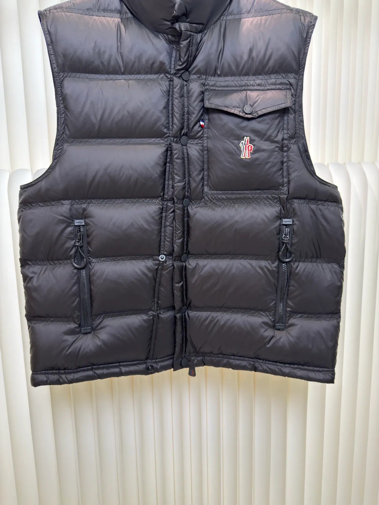 P700 MONCLER masked down vest with reflective markings is made of 90 white duck down with a weight of 130g to ensure excellent thermal insulation performance. After strict screening and processing, the velvet is not easy to get drilled, ensuring durability.  It is made of extremely fine nylon yarn and is woven in a high-density manner. The number of yarns per unit area is large, and the gaps between the yarns are very small, which can effectively block the penetration of cold wind. The fine weave keeps it lightweight and soft to the touch, physically restricting the lightweight down fibers from slipping through the gaps. Nylon fiber itself has good wear resistance and is not easy to snag, which significantly extends its service life and can easily cope with the needs of multiple scenarios.  The lining is made of high-density weaving of 100% polyester fiber, with a thickness of 0.08mm to avoid increasing the overall weight and making it smooth to wear. The high-density weaving structure forms a wind resistance layer to protect the thermal efficiency of the thermal layer from loss. It can withstand the expansion pressure of down and prevent deformation and damage after long-term use.  Through the reflective cloth punching process, the logo is regularly arranged on the triangular reflective mark with dozens of small holes, which not only improves functionality but also enhances recognition and aesthetics. Size 1 2 3 4