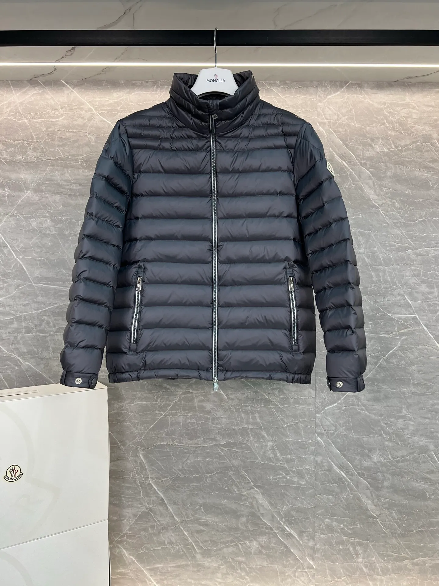 P780 Moncler Arnique short and lightweight men's down jacket. Super soft and delicate fabric, white duck down filling. Extremely lightweight and warm. The Arnique hooded down jacket is made of polyester fiber outer fabric and is Moncler's classic men's design item.  The Arnique short down jacket features classic checkered quilting and signature patch pockets on the sleeves, highlighting the Moncler style. The outer fabric reflects the brand's commitment to lowering its environmental footprint. Modular design, removable hood can be easily put on and taken off according to weather conditions. Details Made of polyester outer fabric Polyester lining Filled with down Size: 1-5 Silver Gray Black