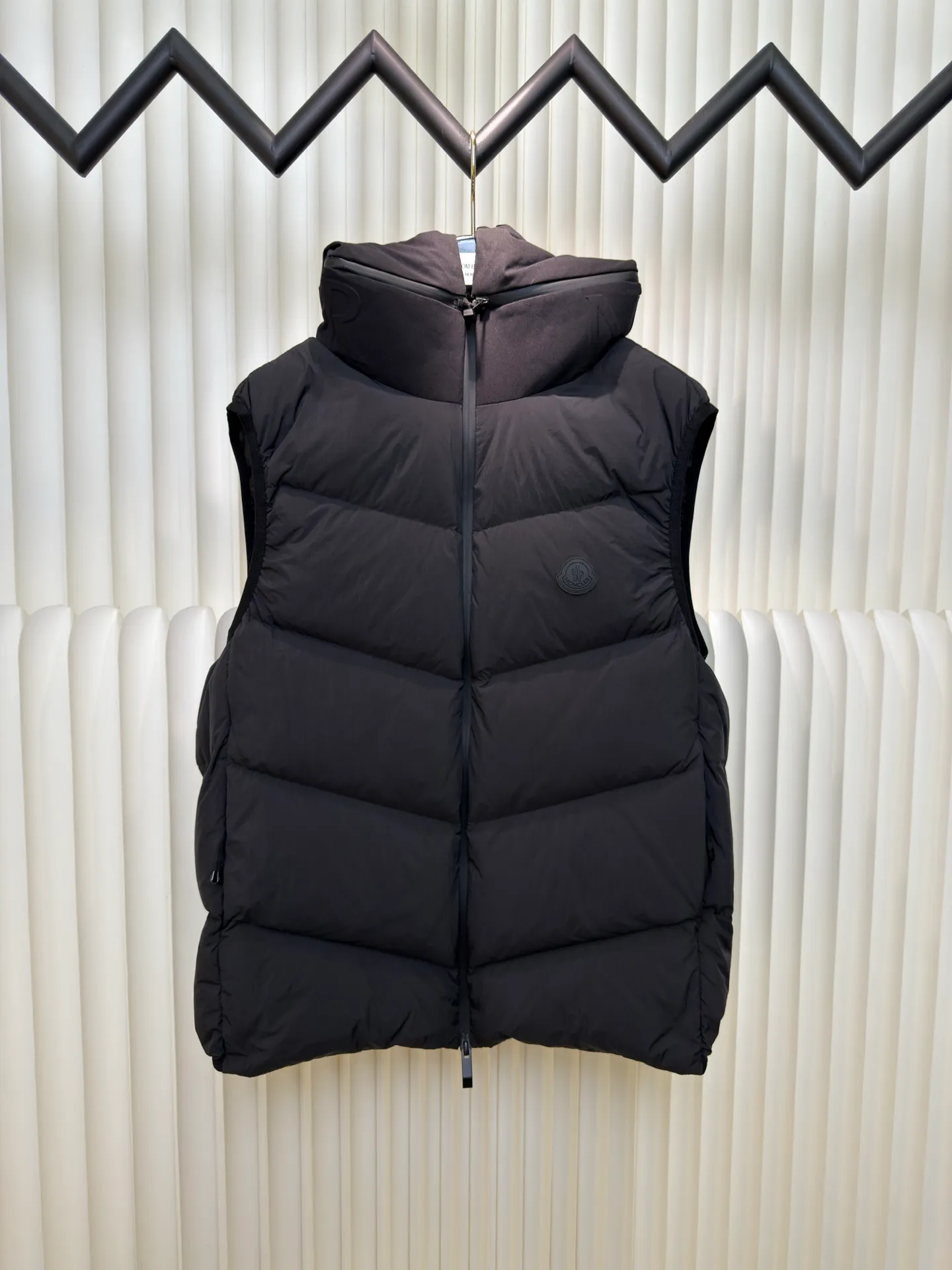P660 MONCLER Badge Hooded Down Vest is made of nylon fabric made of 100% nylon. It is woven into a tight and tough fabric through advanced textile technology. It improves the strength of the frame and effectively blocks the invasion of cold wind and locks in the warm air generated by the down layer. After tight weaving and special treatment, it is waterproof and windproof. The surface forms a certain lotus leaf effect. It is also wrinkle-resistant and not easy to deform, making it easy for daily care.  The quilting stitches are sewn in a diagonal direction at an angle of about 70 degrees, and the diagonal lines are arranged in parallel. The fabric and filling are planned into different functional areas, and the quilting stitches in each partition are relatively independent. This allows the finished product to produce a larger deformation space along the diagonal direction when stretched and pressed, making it less likely to tighten and break, and has rich visual effects, modern and advanced features.  After the hood is removed, the stand-up collar displays an embossed logo of the same color, adding a unique visual experience and tactile details. High-quality filling: It is filled with 90% selected white duck down, weighing 170g, ensuring fluffy rebound, lightness and warmth. Size 1 2 3 4
