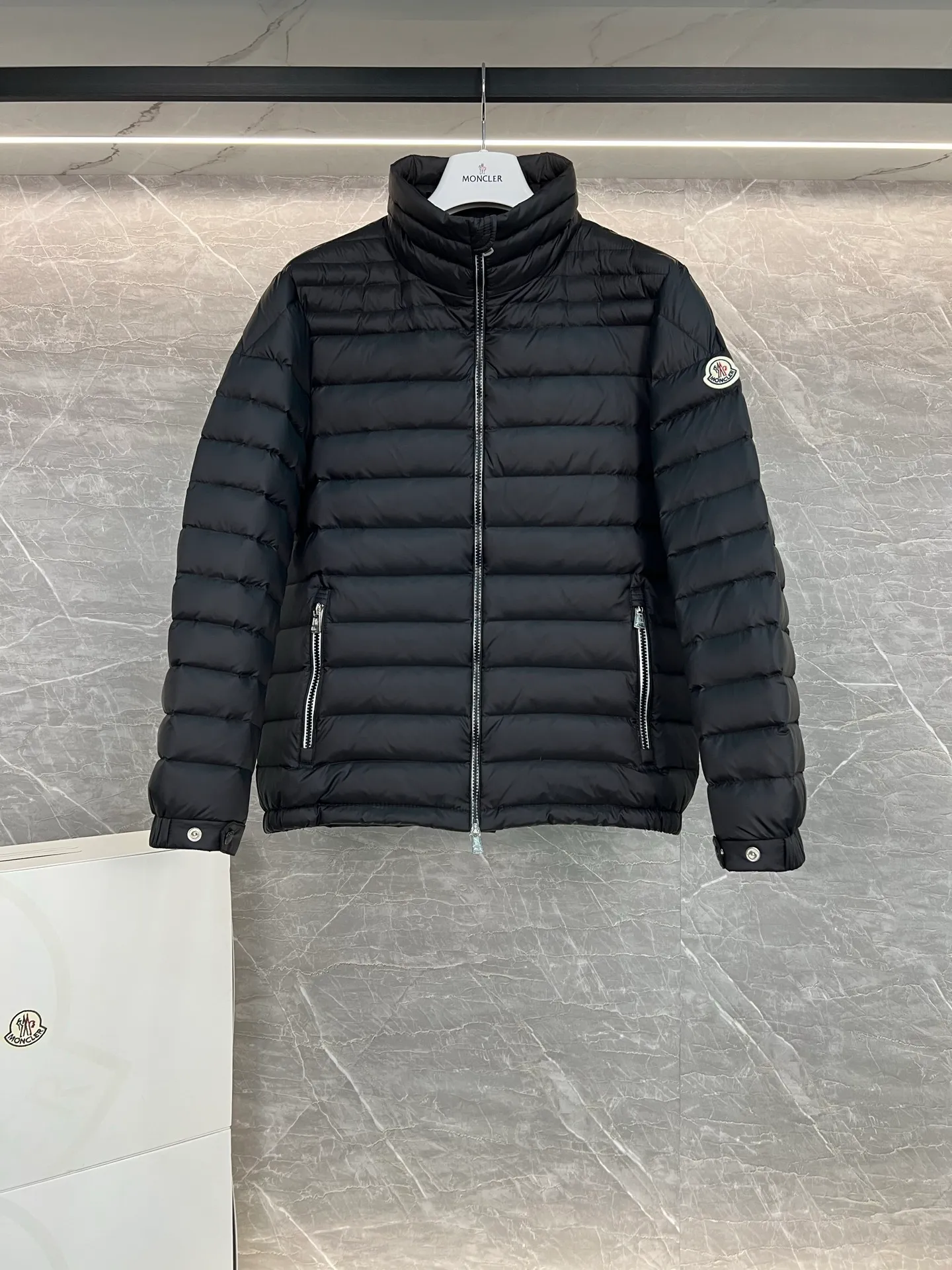 P780 Moncler Arnique short and lightweight men's down jacket. Super soft and delicate fabric, white duck down filling. Extremely lightweight and warm. The Arnique hooded down jacket is made of polyester fiber outer fabric and is Moncler's classic men's design item.  The Arnique short down jacket features classic checkered quilting and signature patch pockets on the sleeves, highlighting the Moncler style. The outer fabric reflects the brand's commitment to lowering its environmental footprint. Modular design, removable hood can be easily put on and taken off according to weather conditions. Details Made of polyester outer fabric Polyester lining Filled with down Size: 1-5 Silver Gray Black