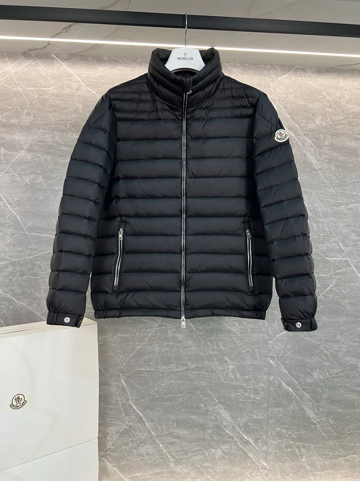 P780 Moncler Arnique short and lightweight men's down jacket. Super soft and delicate fabric, white duck down filling. Extremely lightweight and warm. The Arnique hooded down jacket is made of polyester fiber outer fabric and is Moncler's classic men's design item.  The Arnique short down jacket features classic checkered quilting and signature patch pockets on the sleeves, highlighting the Moncler style. The outer fabric reflects the brand's commitment to lowering its environmental footprint. Modular design, removable hood can be easily put on and taken off according to weather conditions. Details Made of polyester outer fabric Polyester lining Filled with down Size: 1-5 Silver Gray Black