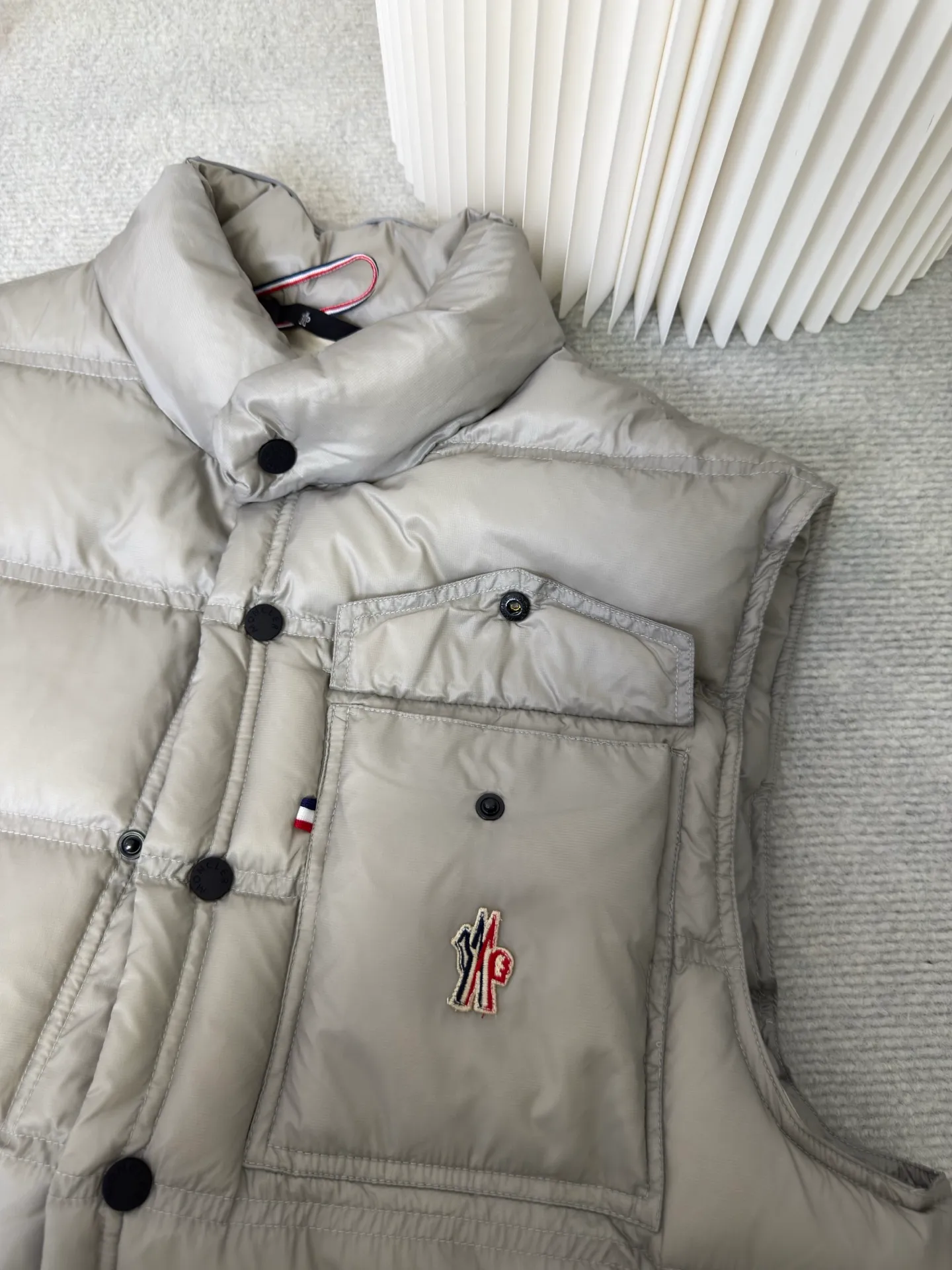 P700 MONCLER masked down vest with reflective markings is made of 90 white duck down with a weight of 130g to ensure excellent thermal insulation performance. After strict screening and processing, the velvet is not easy to get drilled, ensuring durability.  It is made of extremely fine nylon yarn and is woven in a high-density manner. The number of yarns per unit area is large, and the gaps between the yarns are very small, which can effectively block the penetration of cold wind. The fine weave keeps it lightweight and soft to the touch, physically restricting the lightweight down fibers from slipping through the gaps. Nylon fiber itself has good wear resistance and is not easy to snag, which significantly extends its service life and can easily cope with the needs of multiple scenarios.  The lining is made of high-density weaving of 100% polyester fiber, with a thickness of 0.08mm to avoid increasing the overall weight and making it smooth to wear. The high-density weaving structure forms a wind resistance layer to protect the thermal efficiency of the thermal layer from loss. It can withstand the expansion pressure of down and prevent deformation and damage after long-term use.  Through the reflective cloth punching process, the logo is regularly arranged on the triangular reflective mark with dozens of small holes, which not only improves functionality but also enhances recognition and aesthetics. Size 1 2 3 4